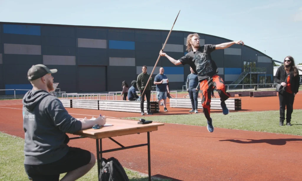 Ung deltager i orange bukser kaster spyd på atletikbanen under STU atletikstævnet, mens tilskuere og personale følger med fra sidelinjen på Langvang Stadion.