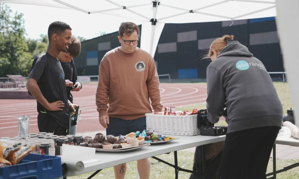 Personale betjener salgsboden under STU atletikstævnet på Langvang Stadion, hvor der sælges forfriskninger og snacks til deltagere og tilskuere.
