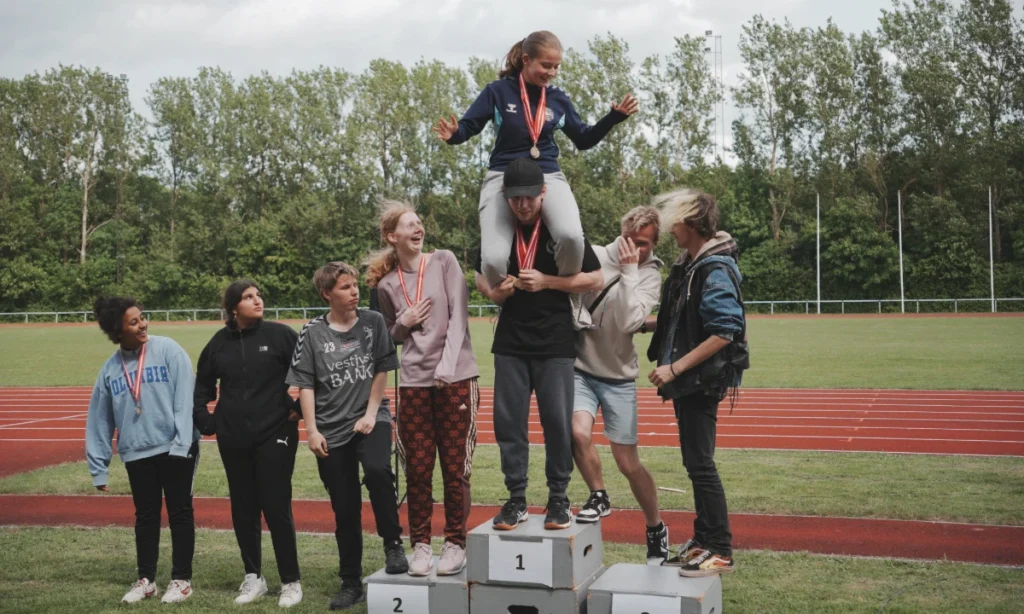 Unge deltagere fejrer sammen på podiet under medaljeoverrækkelsen ved STU atletikstævnet på Langvang Stadion, hvor vindere løftes op af deres medkursister i en spontan hyldest af fællesskab og støtte.