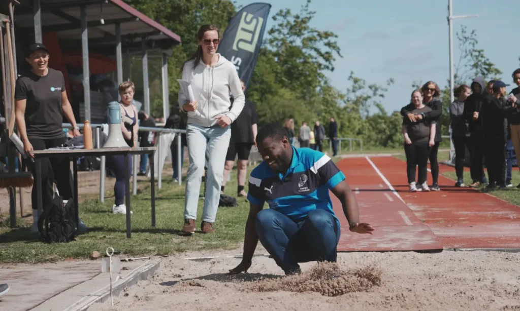 En ung deltager forbereder sig til længdespring på atletikbanen under STU atletikstævnet, mens tilskuere og personale følger med fra sidelinjen.