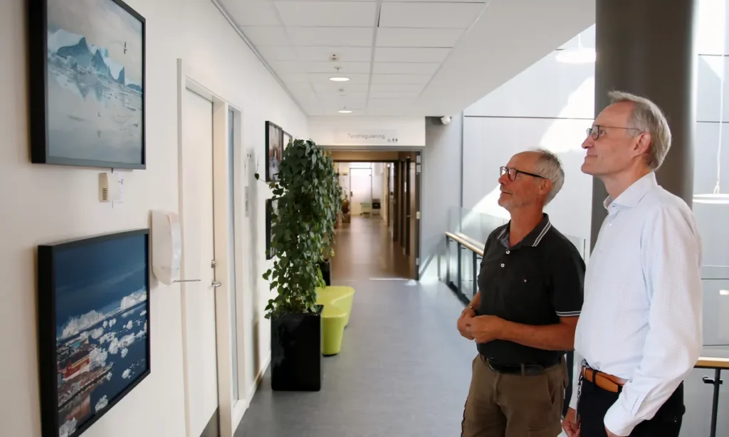 To mænd betragter fotoudstillingen med naturmotiver på hvide vægge i lang korridor med drop-in loft