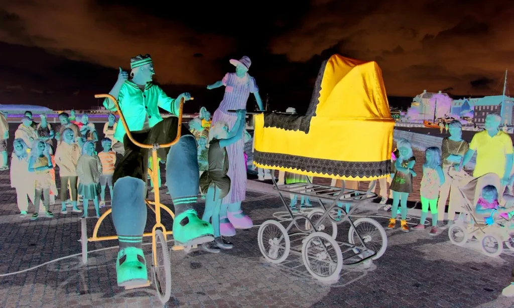 Negativ billede af "Den store familie" med barnevogn med hjul og performere i farverige kostumer omgivet af publikum